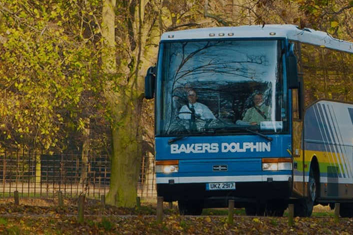 A 'Bakers Dolphin' bus driving on road with scattered leaves and surrounded by autumn trees