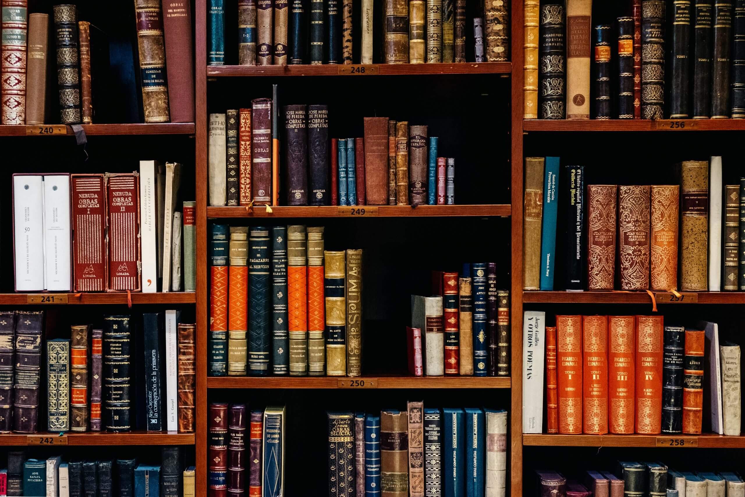 Old beautiful hard back books with colourful, embossed covers are stacked neatly across a unit of chesnut coloured shelves.